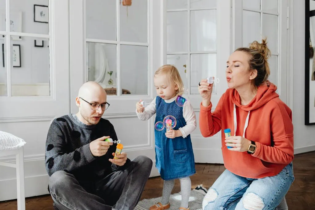 Happy family playing with bubbles indoors — father, mother, and young daughter enjoying quality time together at home.