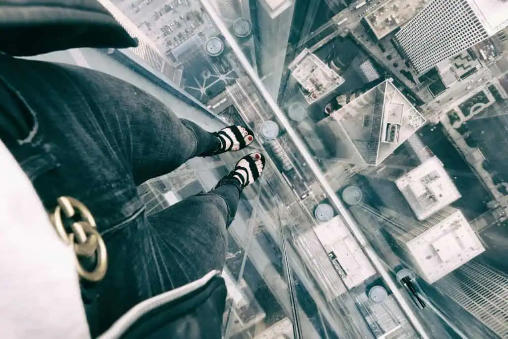 A person standing on a transparent glass floor high above a city, looking down at tall buildings and streets below, illustrating fear of heights.
