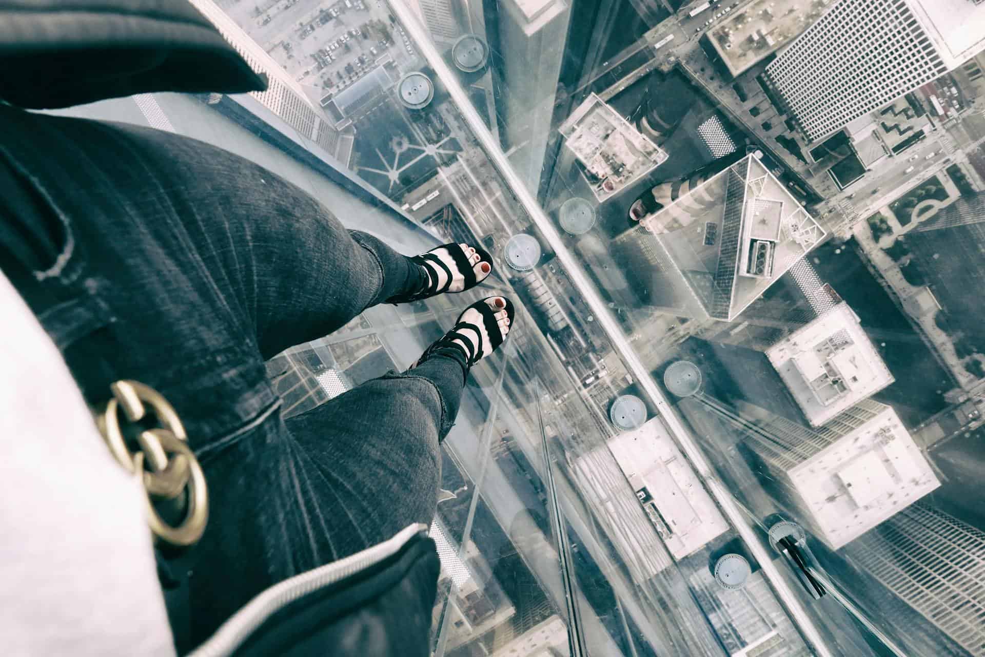 A person standing on a transparent glass floor high above a city, looking down at tall buildings and streets below, illustrating fear of heights.