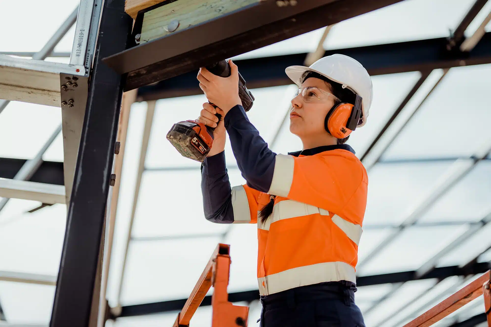 A female construction worker wearing safety gear, including a white helmet, orange high-visibility vest, ear protection, and safety glasses, is using a power drill to work on a steel structure at a construction site.