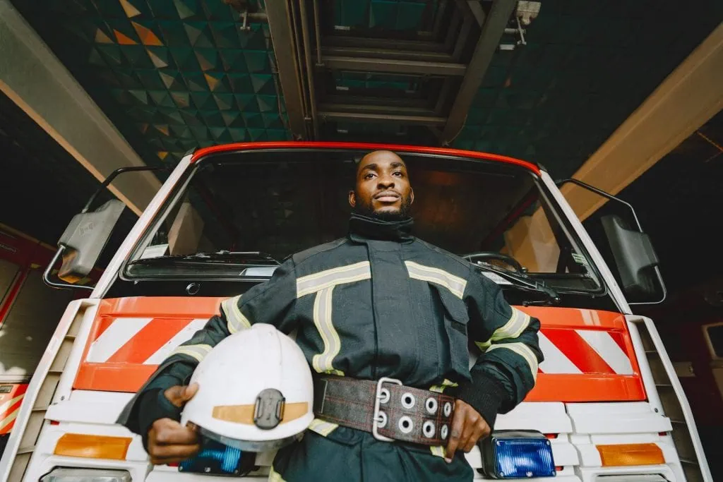 Confident firefighter in uniform standing in front of a fire truck, holding a white helmet.
