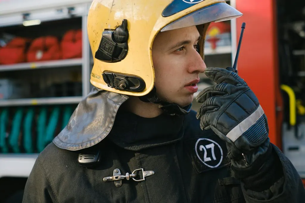 Young firefighter in full gear using a walkie-talkie, standing near a fire truck during emergency response