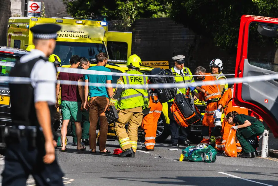 Emergency responders, including firefighters, paramedics, and police officers, attend to a scene involving a red bus and other vehicles on a city street. A group of bystanders, some in shorts and sandals, observes the situation. London Ambulance Service vehicles are present, and the area is cordoned off with police tape. A police officer in uniform stands in the foreground.