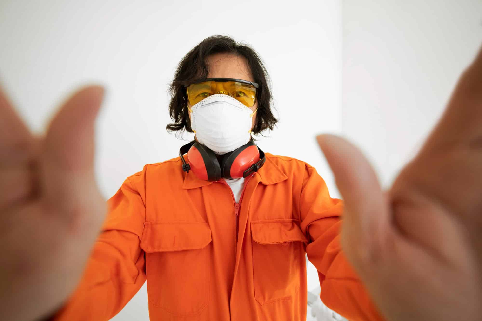 A worker in orange overalls, yellow safety glasses, a white dust mask and red earmuffs around his neck reaches both hands towards the camera