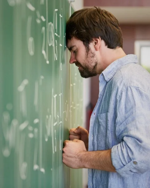 A frustrated man leans his forehead against a chalkboard filled with math equations, clenching his fists in stress or defeat, symbolizing mental exhaustion or academic burnout.