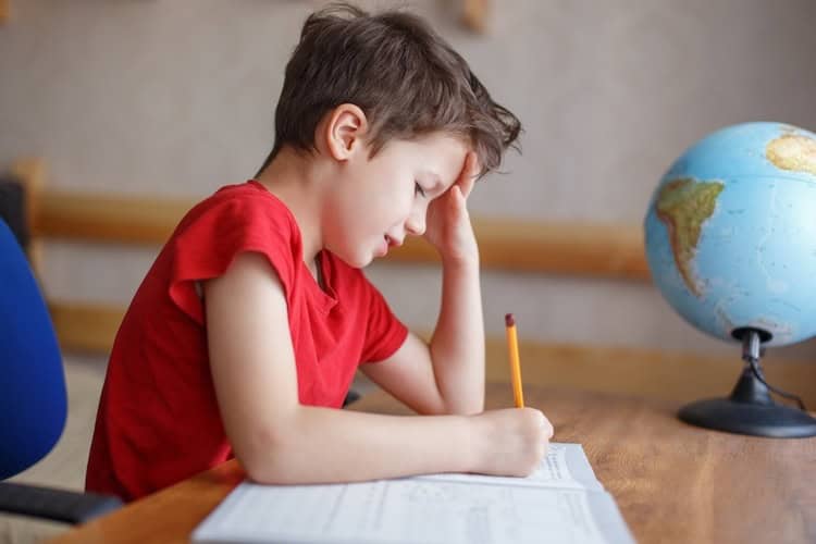 Boy in a red t-shirt at a desk resting his head on one hand while holding a pencil over his schoolwork, with a globe behind him
