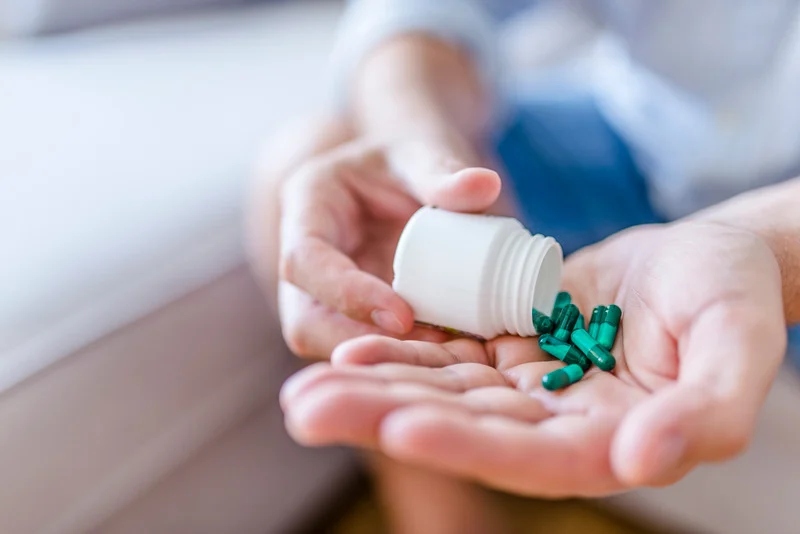 A person pours green capsules from a white medicine bottle into their hand, suggesting medication intake or supplement use, with a soft indoor background.