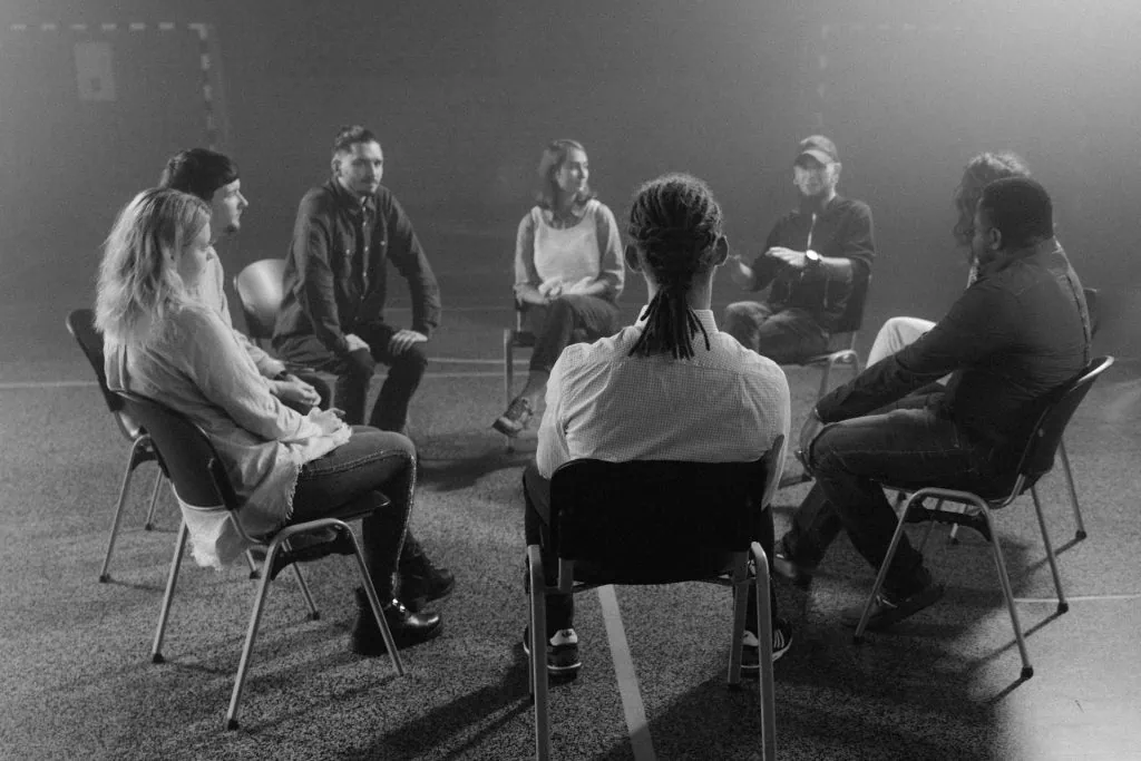 Black and white image of a diverse group of people sitting in a circle during a support or therapy session in a dimly lit room.