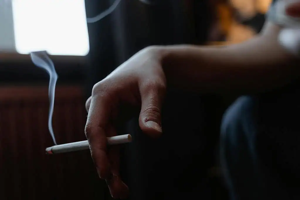 Close-up of a hand holding a lit cigarette indoors with smoke rising