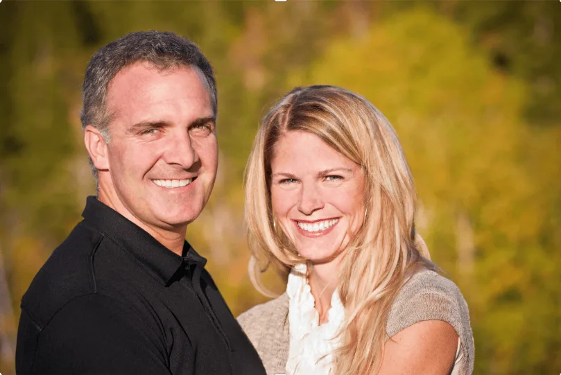 Smiling couple standing close together outdoors with a forest background, both looking at the camera in warm natural light.