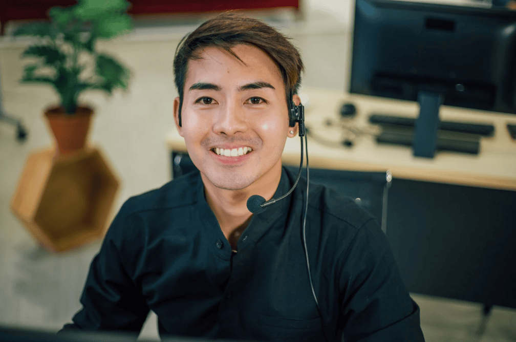 Smiling customer service representative wearing a headset, sitting at a desk in an office.