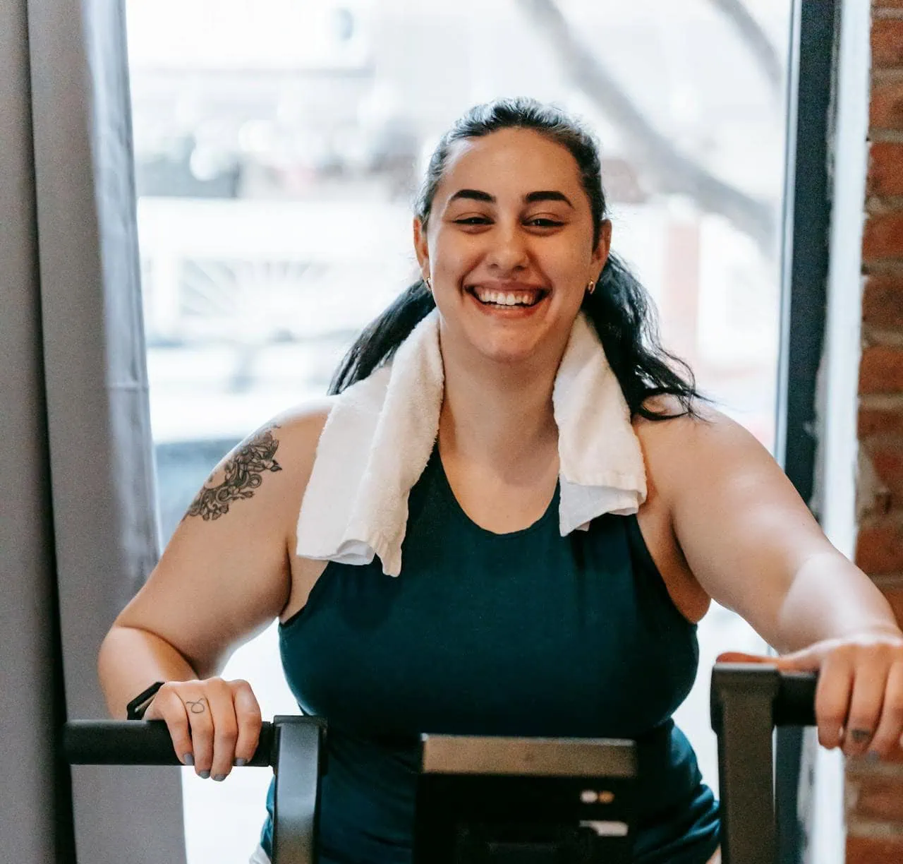 A smiling woman with a white towel around her neck is exercising on a stationary bike indoors. She is wearing a dark teal tank top and light-colored pants, with a tattoo visible on her upper arm.