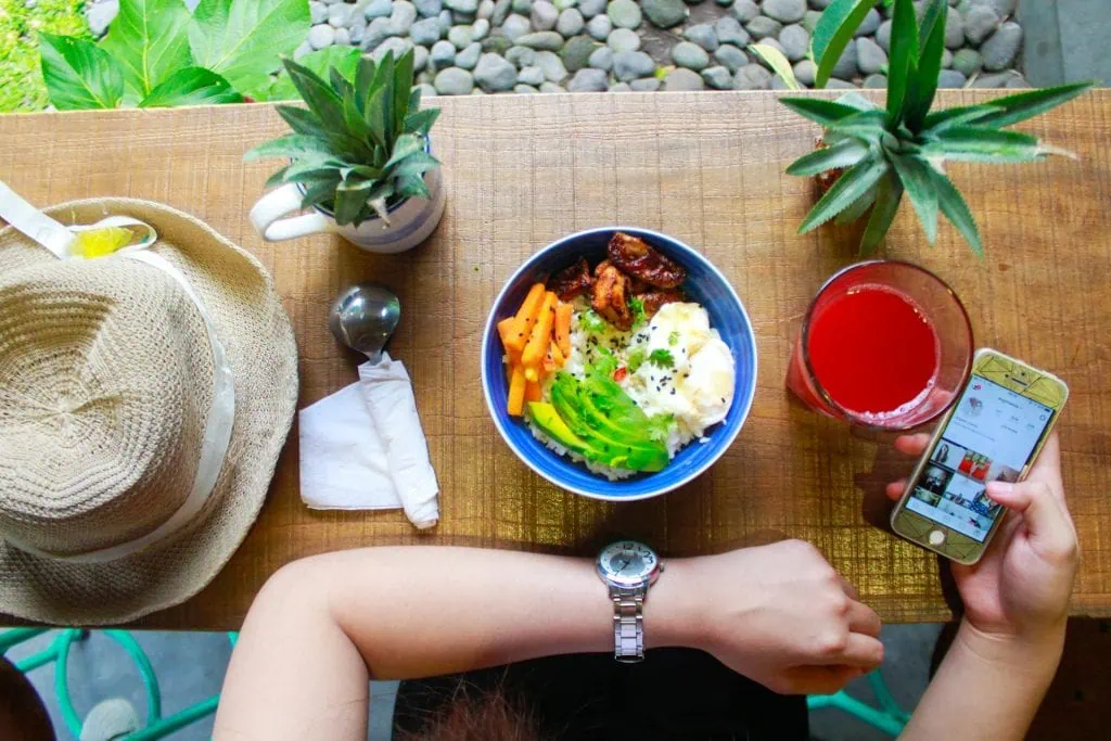 Healthy meal with rice, grilled chicken, avocado, carrots, and a glass of juice, with a person checking their phone