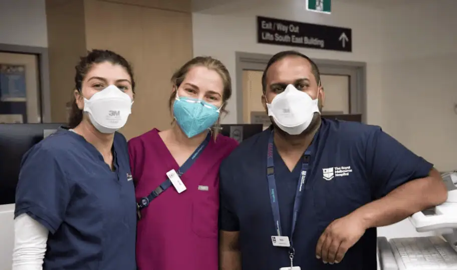 Three healthcare workers in a hospital setting wearing face masks, scrubs, and ID badges, smiling at the camera.
