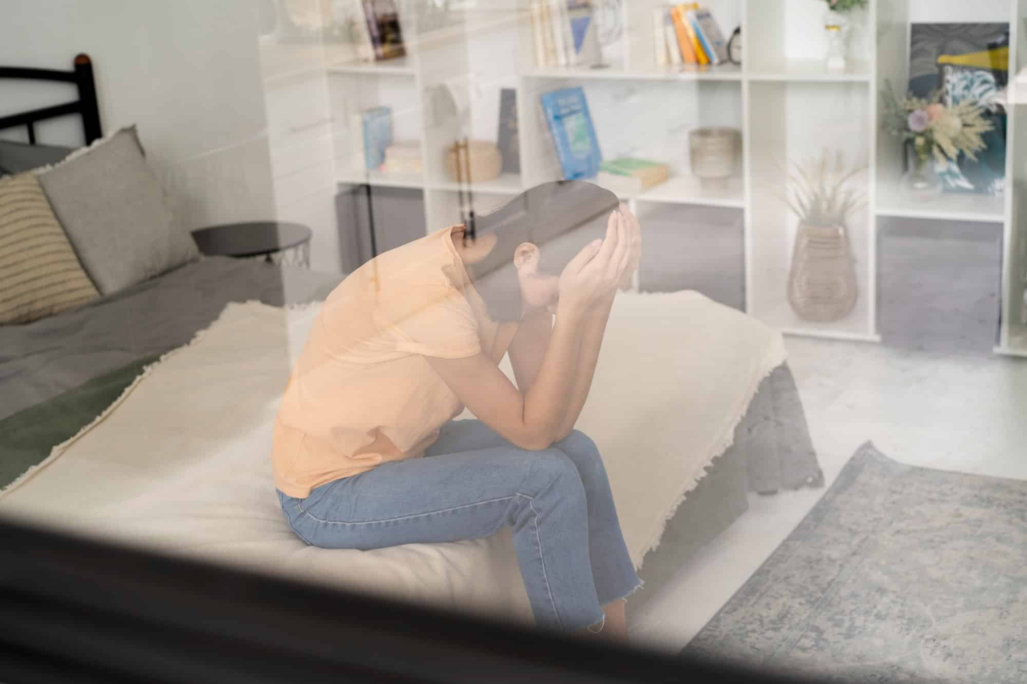 Woman sitting on the edge of a bed holding her head in both hands, seen through a window