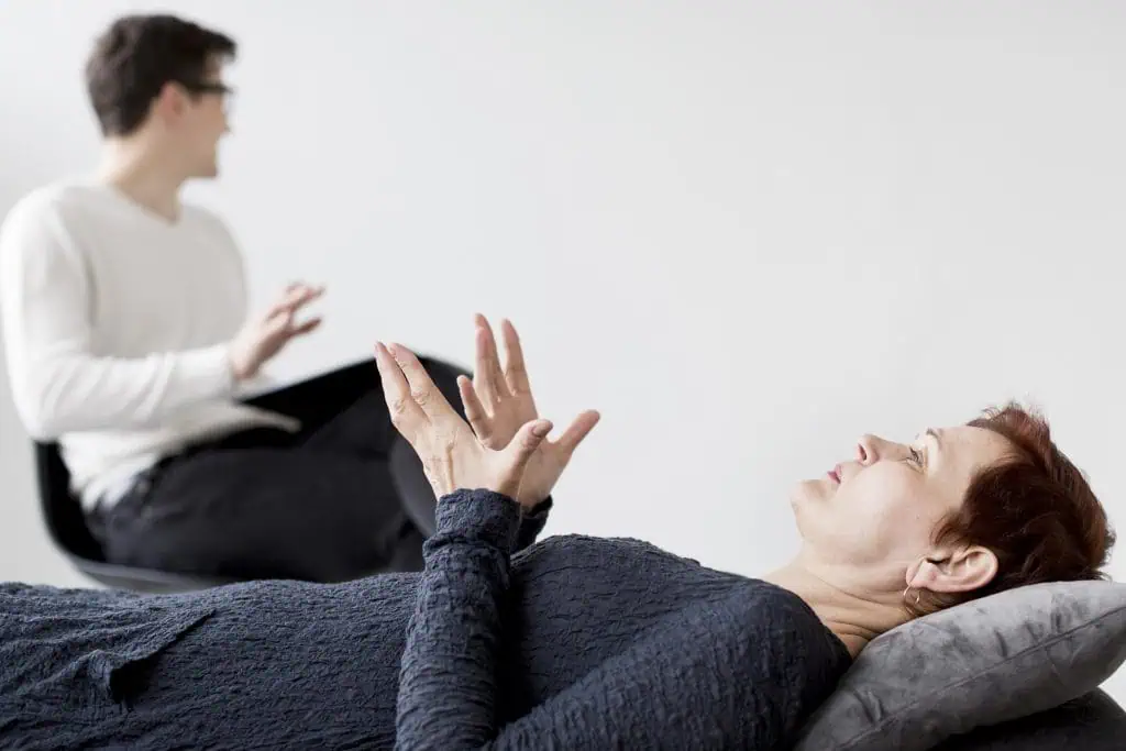 A woman lying on a therapy couch talking to a male psychologist seated nearby, during a counseling or hypnotherapy session in a bright room.