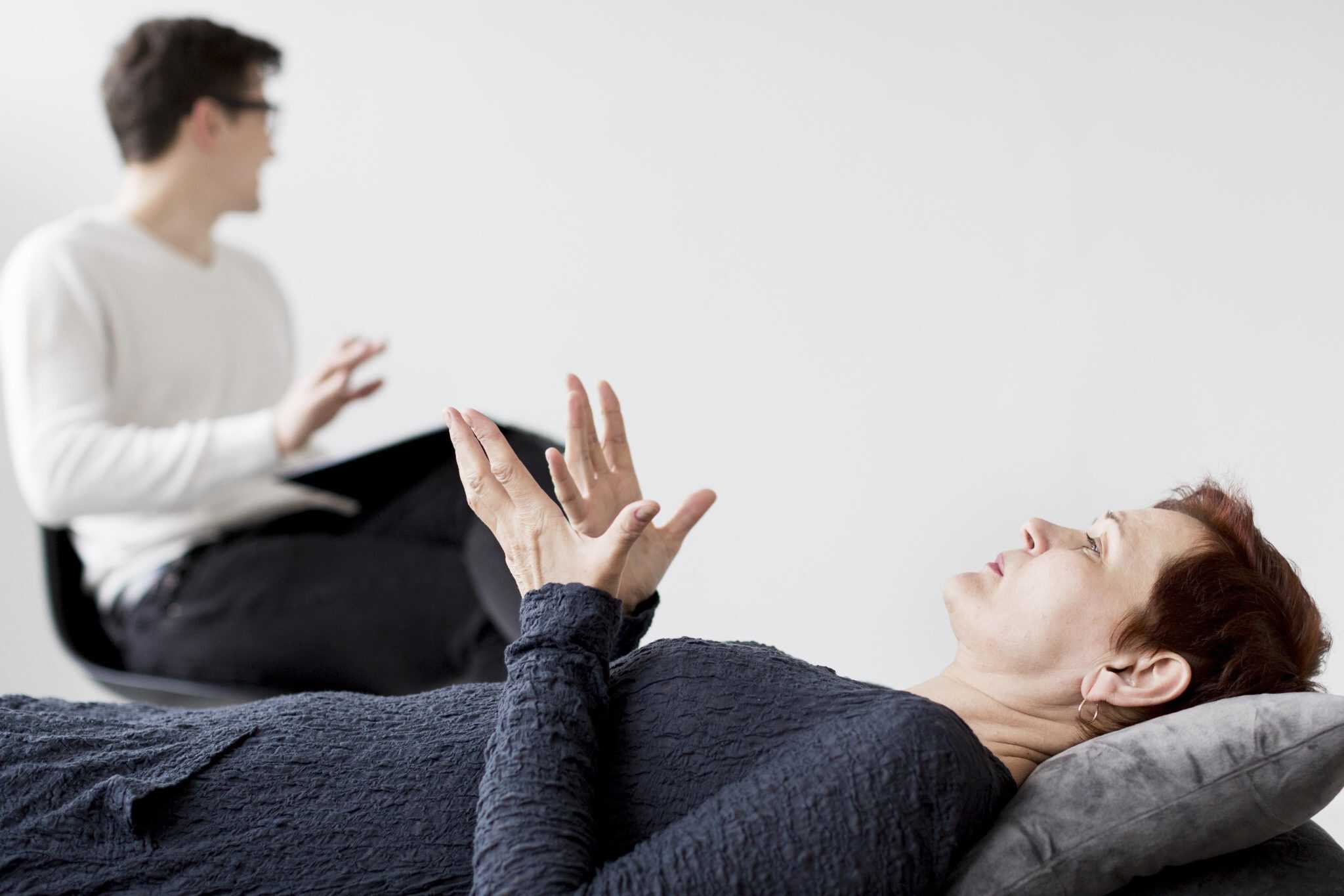 A woman lying on a therapy couch talking to a male psychologist seated nearby, during a counseling or hypnotherapy session in a bright room.