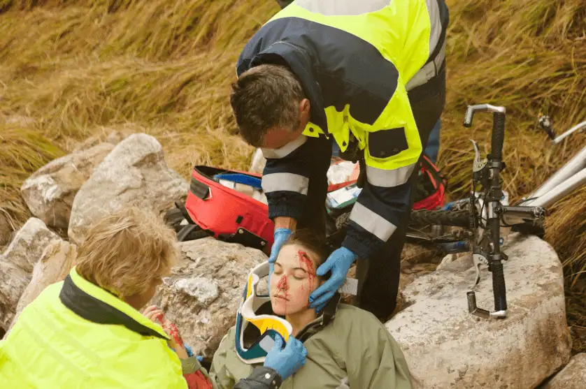 Emergency responders in high-visibility jackets provide medical assistance to an injured cyclist lying on rocky terrain. The patient has a facial wound and is being fitted with a cervical collar. A damaged bicycle and medical supplies are visible in the background.