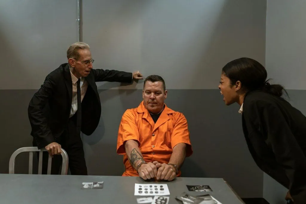 An inmate sits in a dimly lit interrogation room, showing visible signs of stress or anxiety, possibly during questioning by authorities.