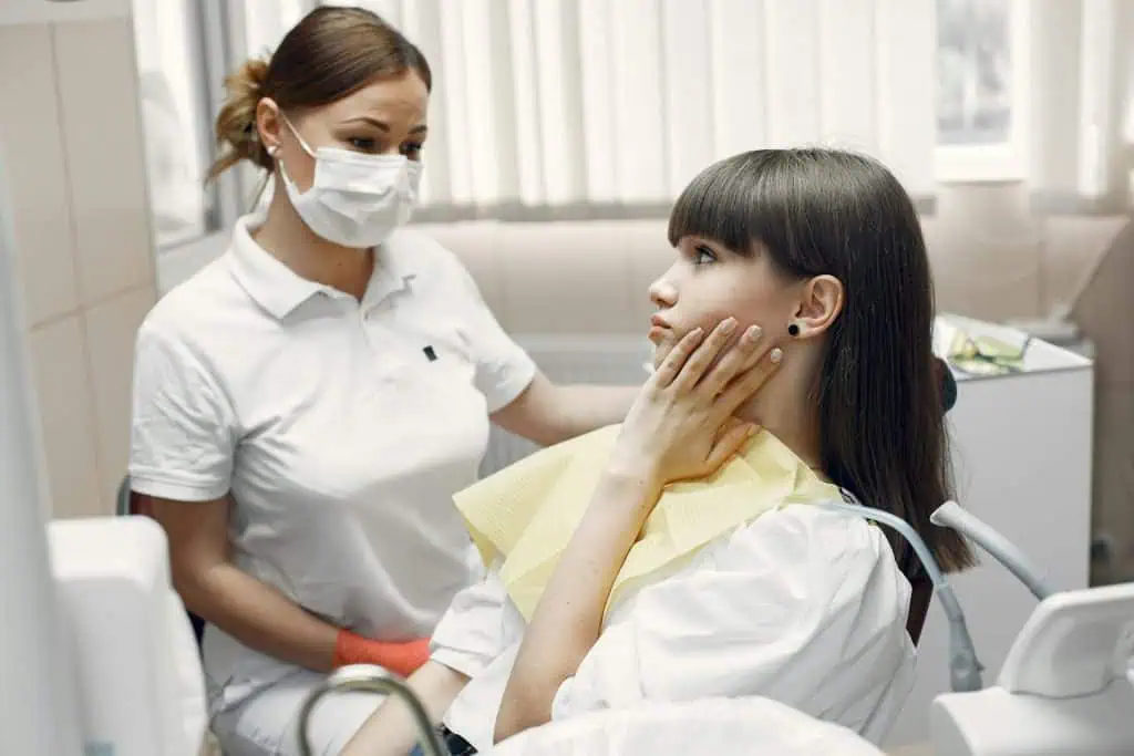 A young woman sitting in a dental chair holding her jaw in discomfort while speaking with a female dentist wearing a mask and gloves in a bright dental clinic.