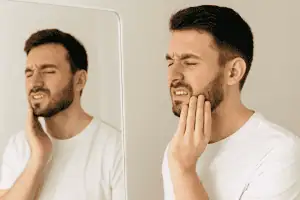 A young man in a white shirt stands in front of a mirror, holding his jaw with a pained expression, showing visible discomfort from jaw pain or teeth grinding.