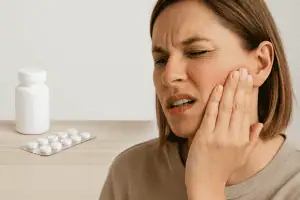 Woman experiencing jaw pain, pressing her hand to the side of her face, with a pill bottle and blister pack of tablets on a wooden table in the foreground, representing treatment options for TMJ pain or bruxism.