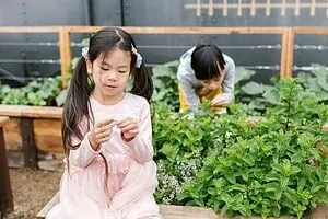 Two children exploring a garden, with one girl calmly observing a plant and another child picking herbs in the background—an example of mindful outdoor activity for kids with ADHD.