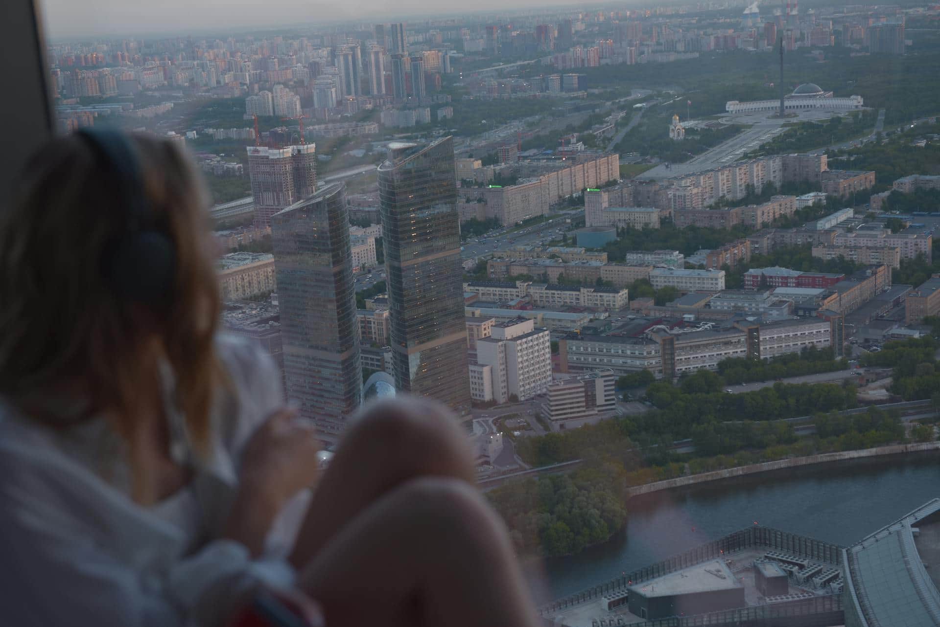 A woman with headphones on, blurred in the foreground, looking out a high window over a city at dusk.