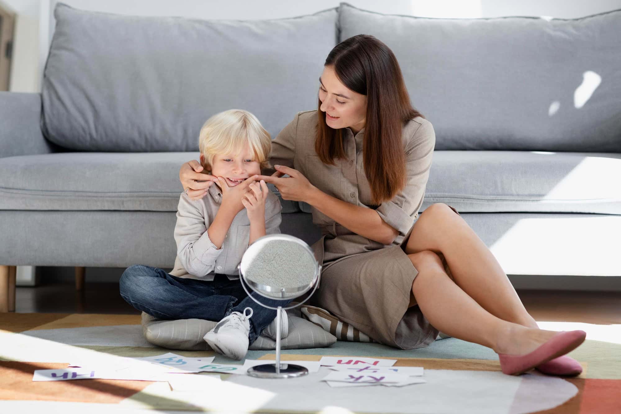 A woman and a young blond boy sitting on the floor in front of a grey sofa, both holding the boy's cheeks and mouth while looking at a small mirror, with letter cards on the rug.