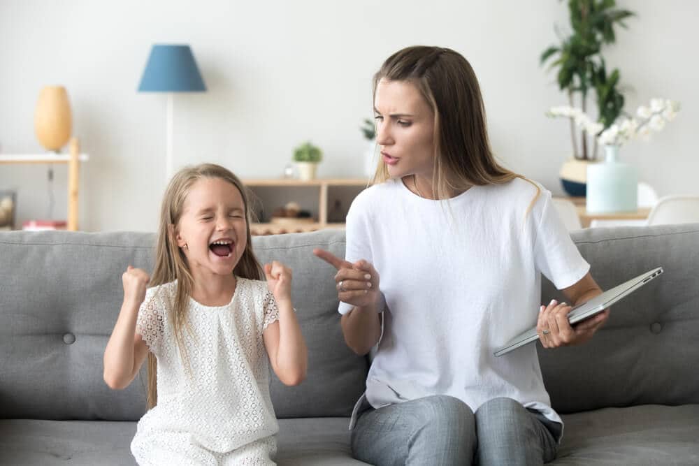 Young girl with her fists raised and her mouth open, looking upset, facing a woman who holds a tablet on a grey sofa