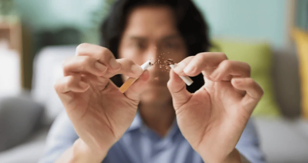 A man in a blue shirt holds a cigarette with both hands and snaps it in half, symbolizing quitting smoking.