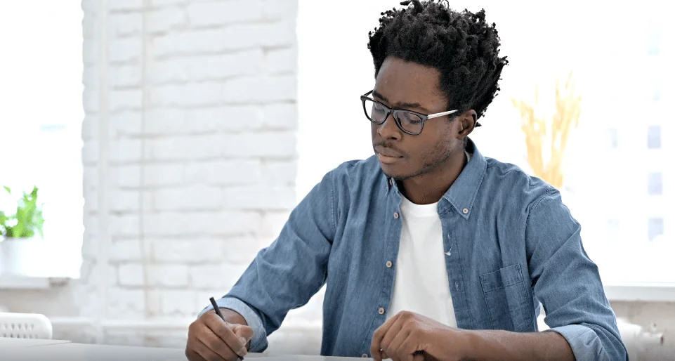 Focused man in glasses writing in a notebook at a desk, possibly journaling or working through thoughts.