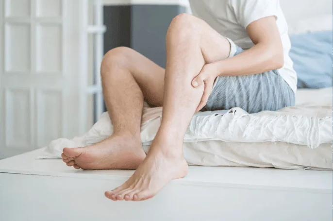 A man sitting on the edge of his bed, holding his calf, possibly experiencing leg cramps or muscle pain in the morning. The bright bedroom setting suggests early hours.