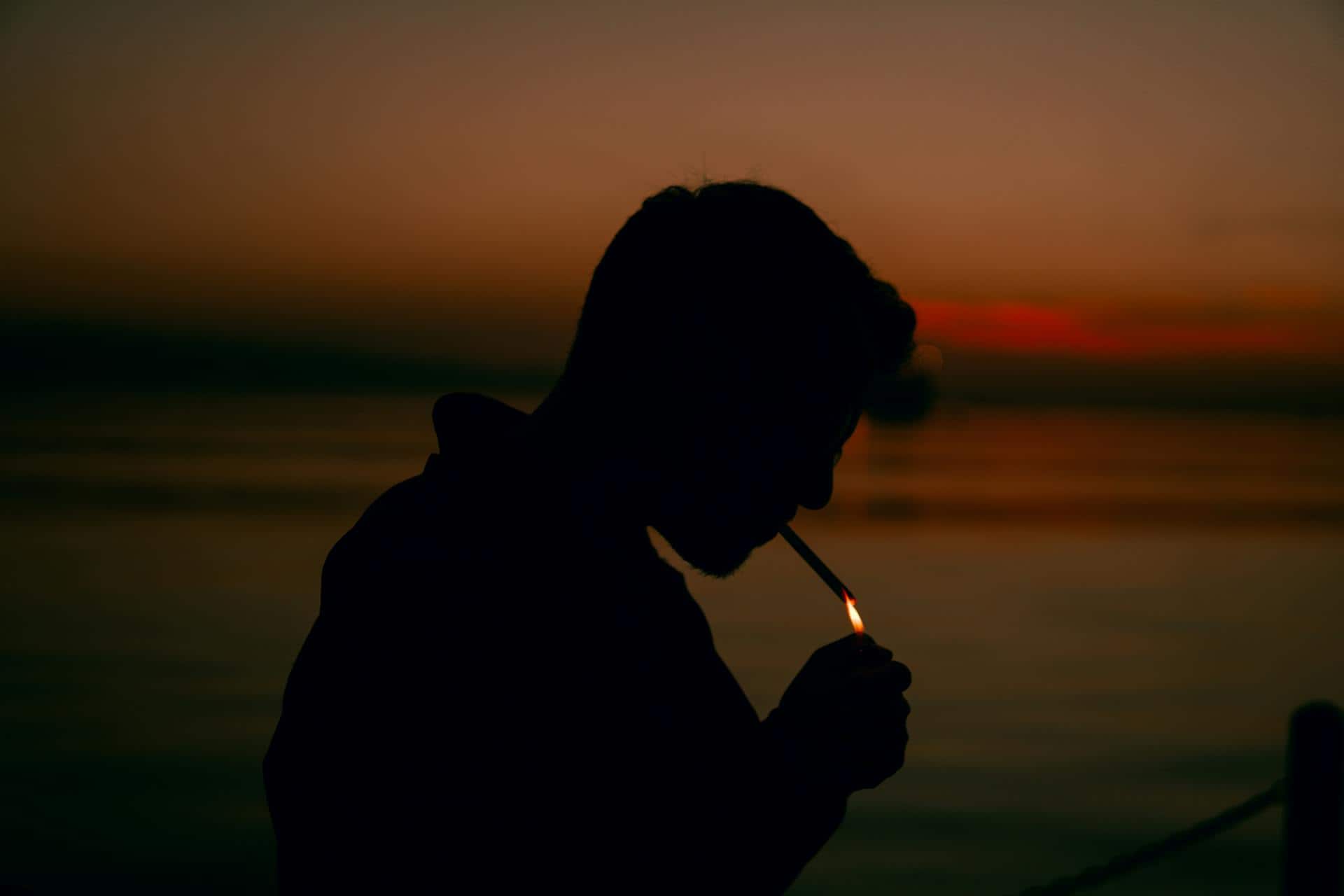 Silhouette of a bearded man lighting a cigarette with a lighter against an orange dusk sky over water