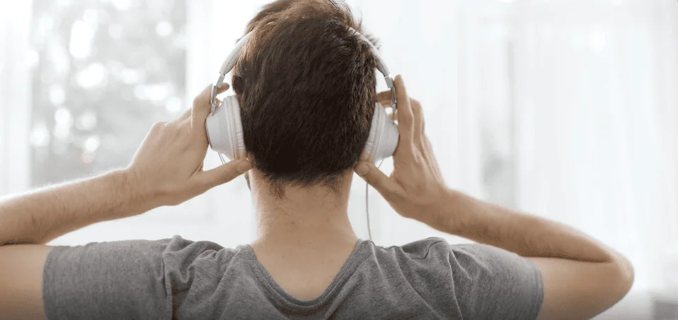 Back view of a man wearing a gray shirt putting on white headphones, possibly preparing to listen to music or an audio session.