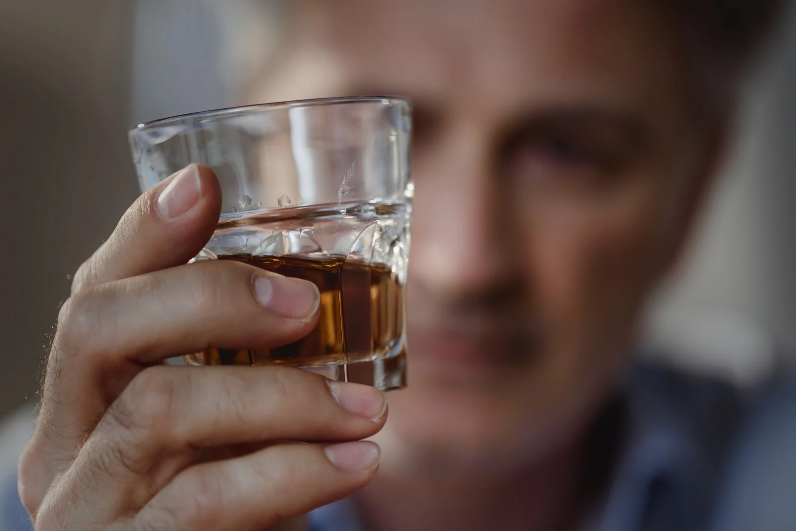 A man holds a glass of amber-colored liquor in focus, while his face remains blurred in the background, capturing a moment of deep reflection or inner conflict related to alcohol consumption.