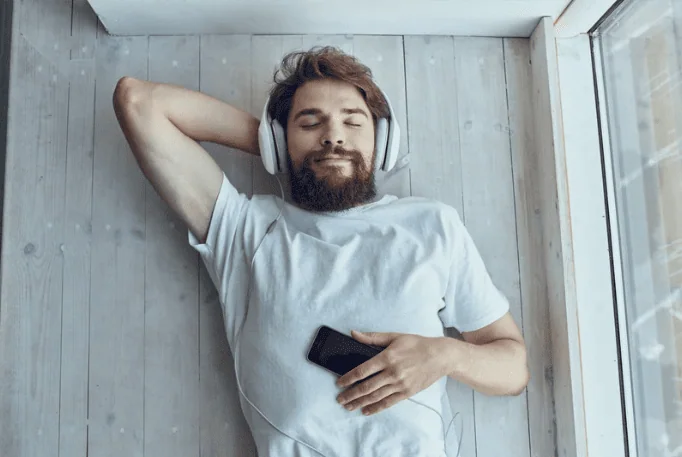 Calm woman leaning back on a couch with eyes closed, enjoying audio through white headphones in a bright room.