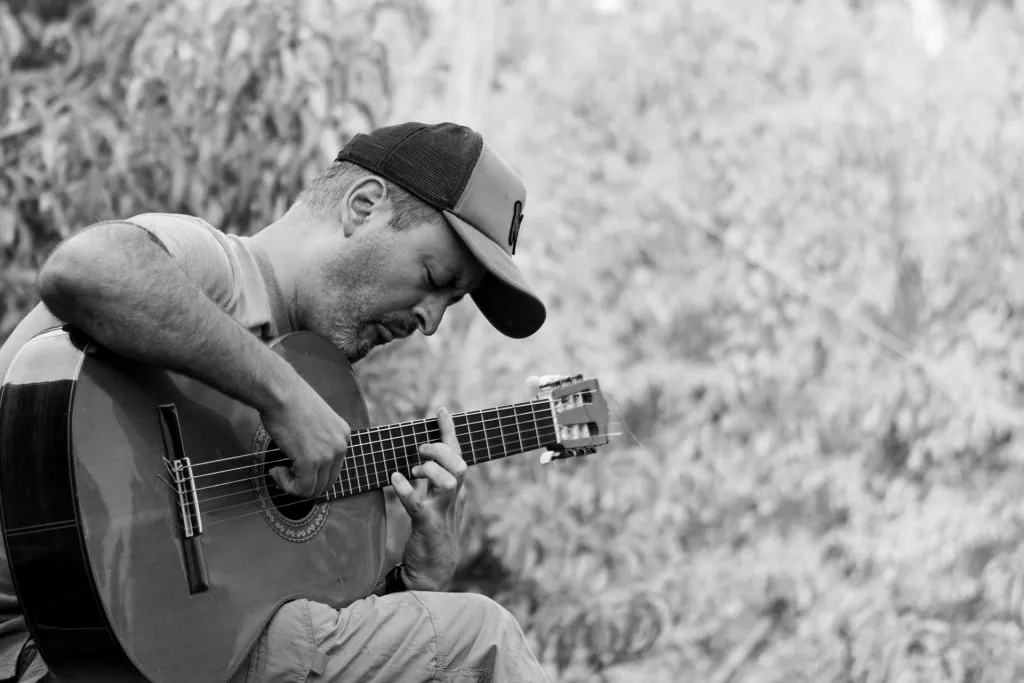 Black and white photo of a man wearing a cap, deeply focused while playing an acoustic guitar outdoors.
