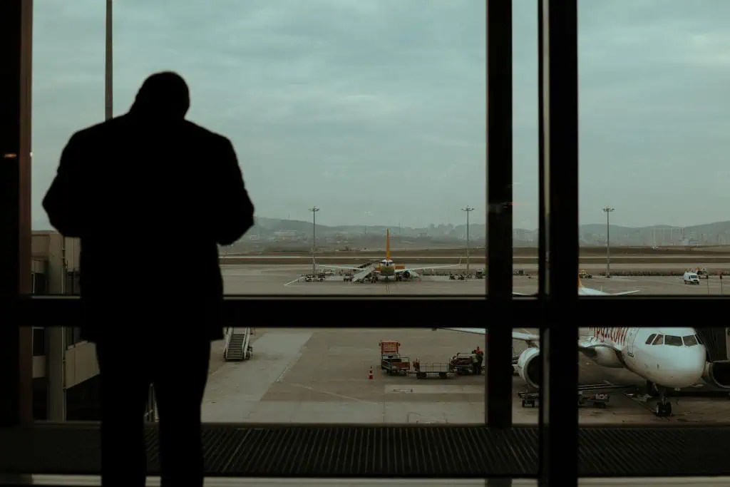 Silhouette of a man standing in an airport terminal, looking out at airplanes on the tarmac under a cloudy sky, symbolizing contemplation or anxiety before flying.