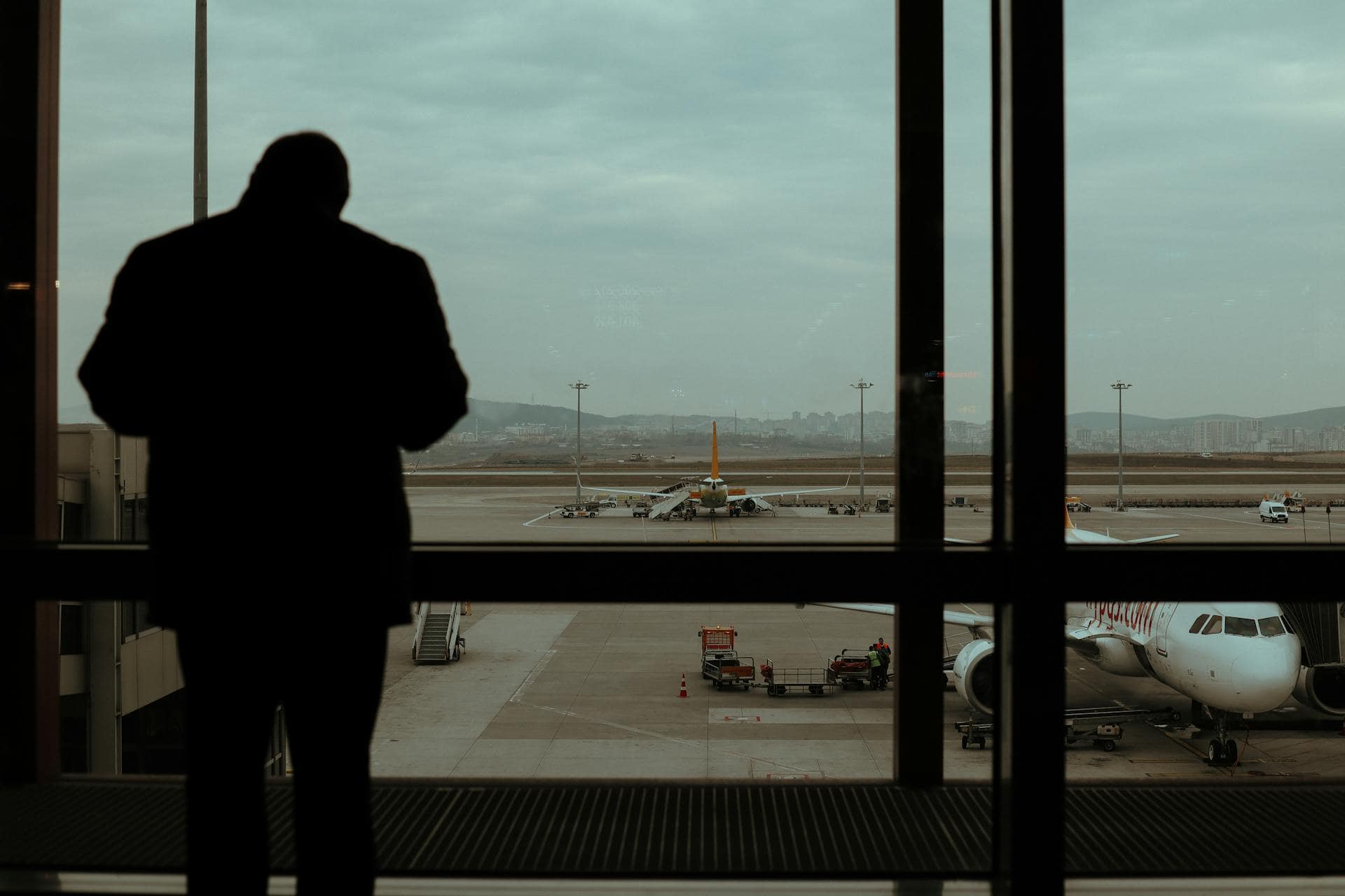 Silhouette of a man standing in an airport terminal, looking out at airplanes on the tarmac under a cloudy sky, symbolizing contemplation or anxiety before flying.