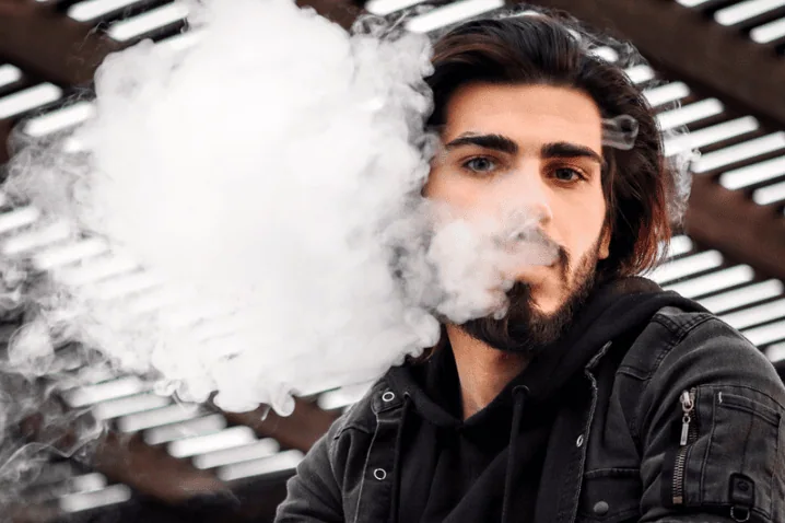 A bearded man in a black hoodie exhales a large cloud of vape smoke outdoors under a shaded structure.