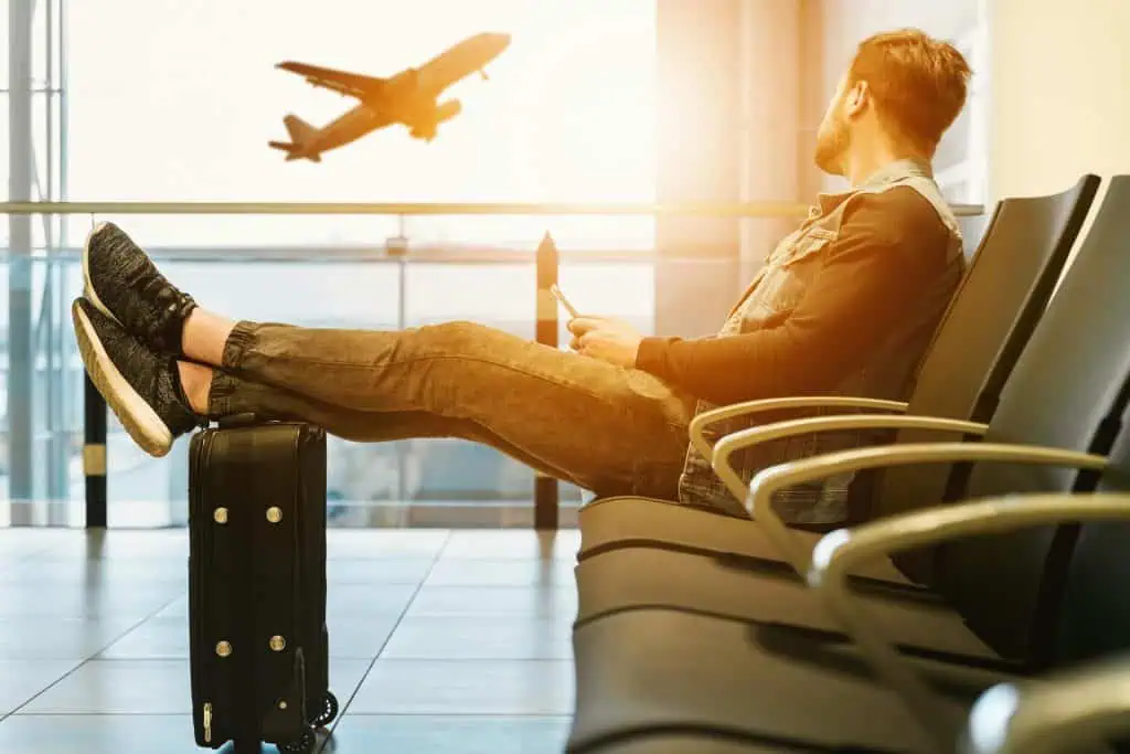 Man sitting in an airport lounge with legs on his suitcase, looking out the window as a plane takes off, capturing anticipation and reflective emotions before flying.