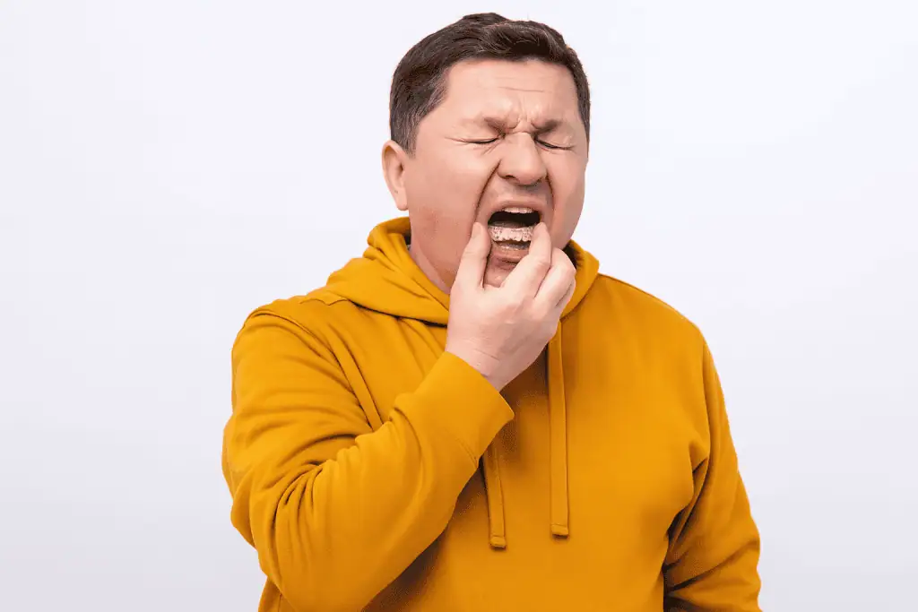 A middle-aged man in a mustard hoodie placing a mouthguard in his mouth with a pained expression, standing against a plain white background.