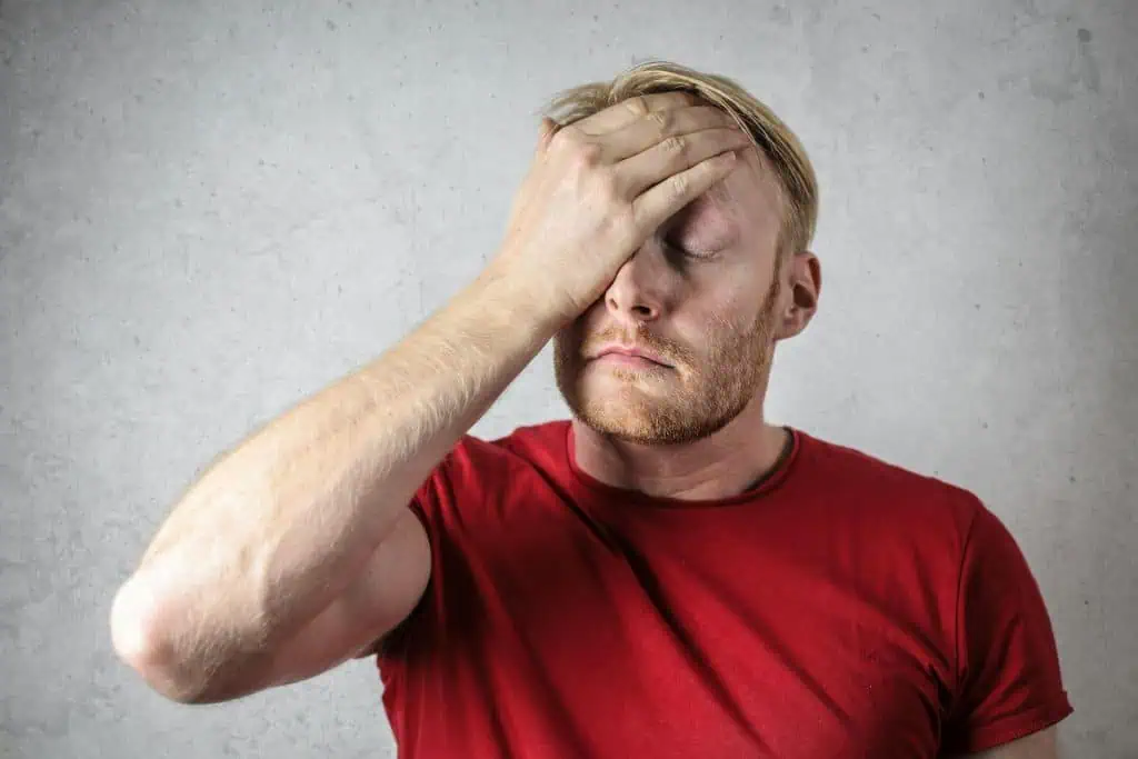 Man in a red shirt with eyes closed, holding his forehead in apparent stress or pain indicating one of the causes of bruxism.