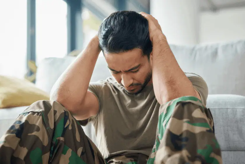 A distressed military veteran sits on the floor with his back against a couch, gripping his head in frustration. He wears a camouflage uniform and an olive-green shirt, conveying a sense of emotional struggle.