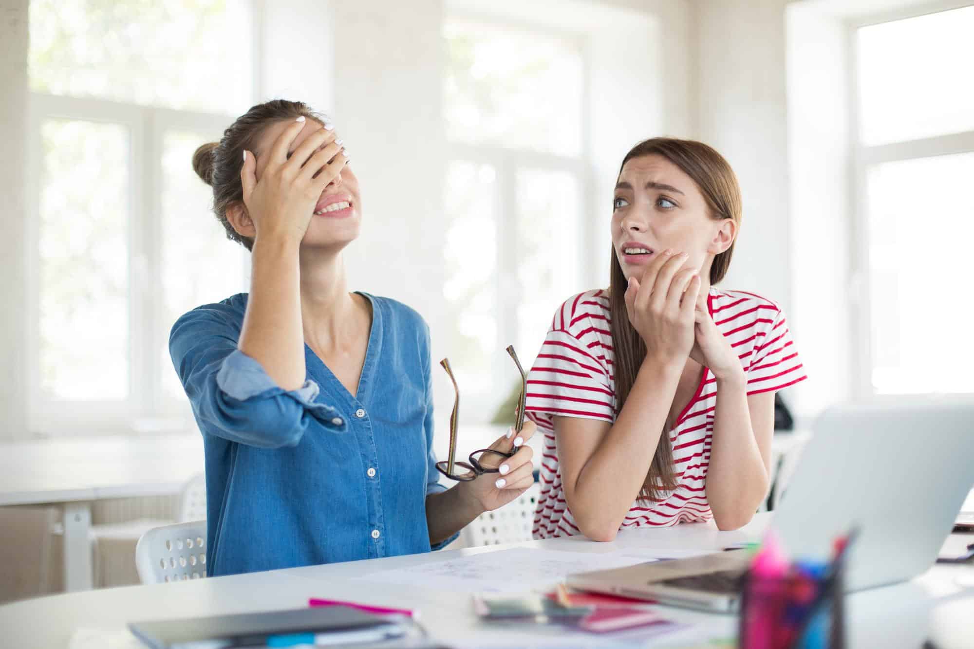 Two women at a desk in a bright office, one covering her eyes with her hand and the other grimacing with her hands at her chin.