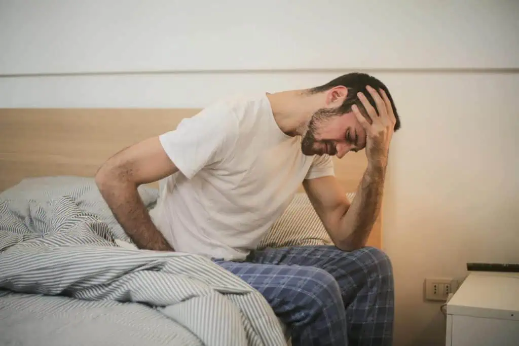 Man sitting on bed holding his head in pain, possibly experiencing a morning headache from sleep bruxism.