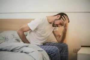 Man sitting on bed holding his head in pain, possibly experiencing a morning headache from sleep bruxism.