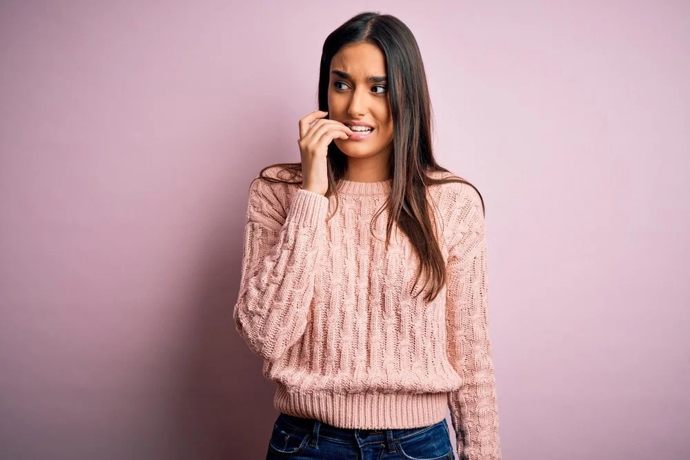 A young woman with long dark hair wearing a light pink knitted sweater and jeans stands against a pink background, nervously biting her fingernail and looking to the side.
