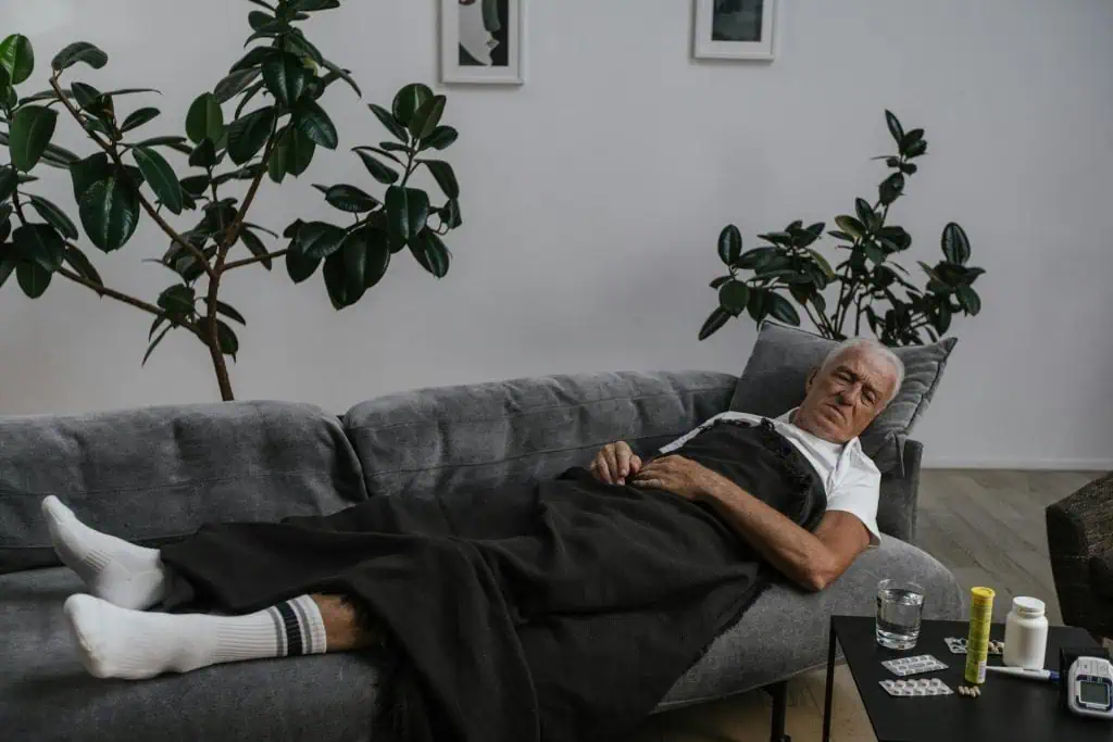 Elderly man lying on a couch under a blanket, surrounded by medication, possibly recovering from sleep-related discomfort or chronic pain.